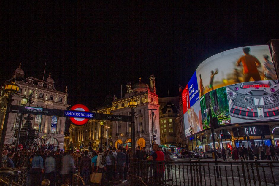 Nighttime scene of an underground subway entrance with a round, illuminated red and white London Underground sign and a blue rectangular 'UNDERGROUND' sign attached to a black metal frame. To the righ