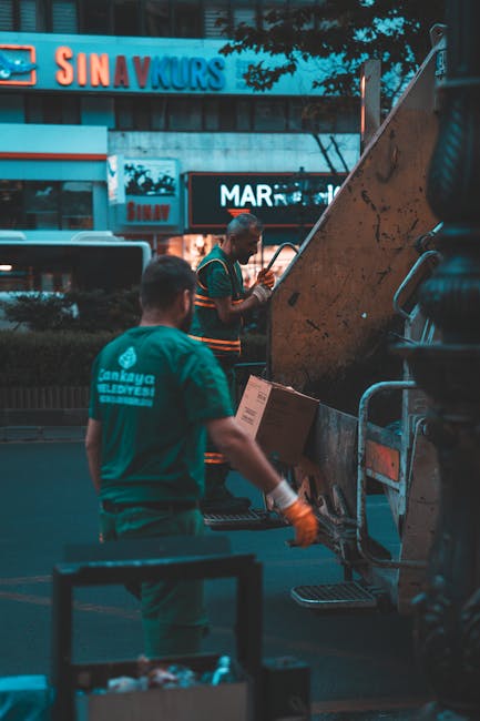 Two men are engaged in rubbish collection outdoors on a paved area, with one wearing yellow gloves and black clothing, and the other dressed in a grey T-shirt and dark trousers. They are lifting and t