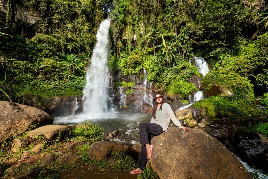 A woman with long brown hair wearing sunglasses, a light grey long-sleeved top, dark trousers, and brown sandals, is seated on a large, smooth boulder in front of a lush, green rainforest scene. Behin